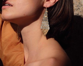 Può includere: Una donna con un top marrone e capelli lunghi e scuri. Indossa orecchini lunghi geometrici dorati e argentati.