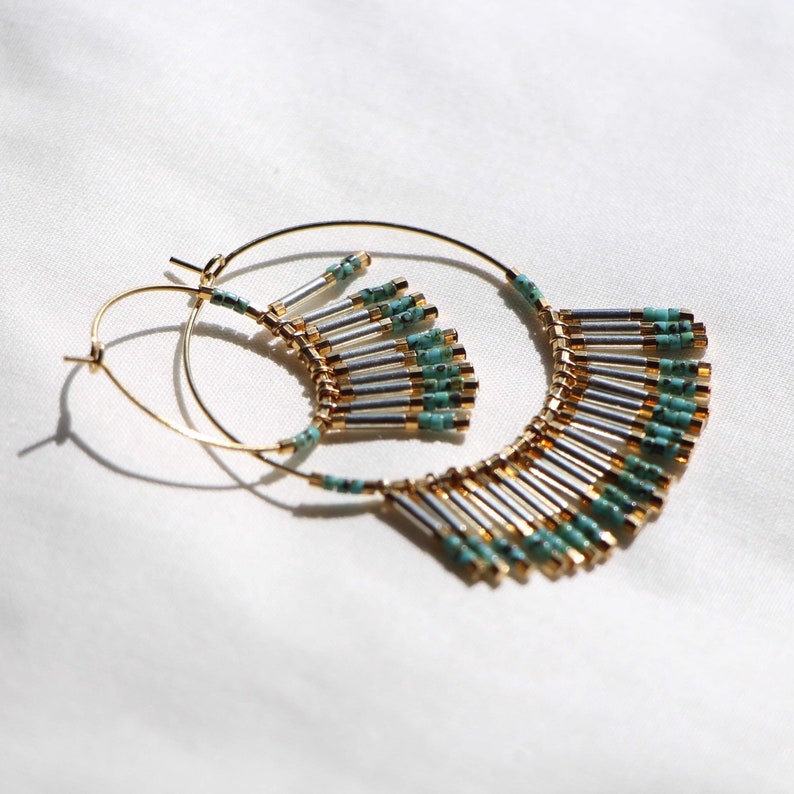 May include: Gold hoop earrings with a turquoise and silver beaded fringe. The fringe is made of small, cylindrical beads that are strung together in a cascading pattern.