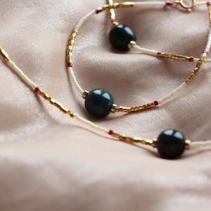 May include: Two necklaces with gold and white beads and dark green round beads. The necklaces are on a light brown fabric background.