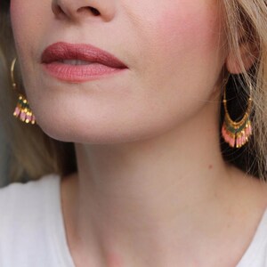 May include: A close-up of a person's face wearing gold hoop earrings with pink and green beaded fringe. The earrings are dangling from the person's ears.