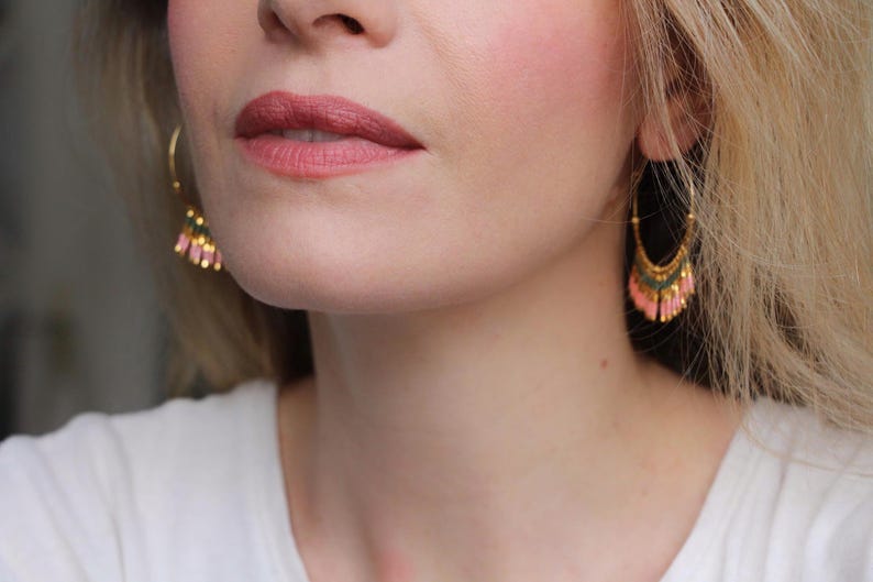 May include: A close-up of a person's face wearing gold hoop earrings with pink and green beaded fringe. The earrings are dangling from the person's ears.