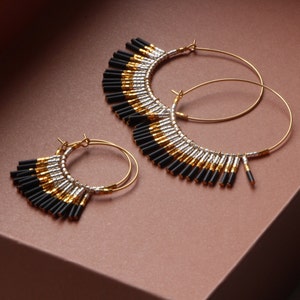 May include: A pair of gold hoop earrings with black and silver beads hanging from the bottom. The earrings are on a brown surface.