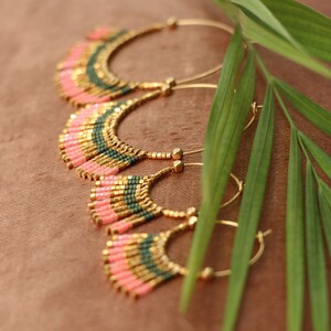 May include: Three pairs of gold hoop earrings with pink and green beaded fringe. The earrings are arranged in a cascading pattern, with the largest pair at the top and the smallest pair at the bottom.