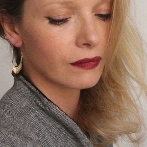 May include: A woman with long blonde hair wearing a black and white patterned jacket and gold hoop earrings with black tassels.