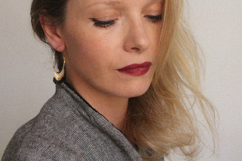 May include: A woman with long blonde hair wearing a black and white patterned jacket and gold hoop earrings with black tassels.