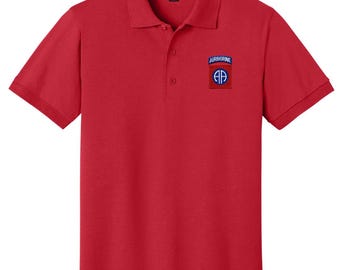Puede incluir: Un polo rojo con mangas cortas y cuello. La camisa tiene un pequeño parche en el pecho con la palabra "AIRBORNE" encima de un emblema azul y blanco. La camisa tiene una tapeta de dos botones.
