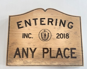 Op de afbeelding: Een houten bord met de tekst "ENTERING INC. 2018 ANY PLACE" in zwarte letters. Het bord heeft een afgeronde bovenkant en een donkerbruine rand. Het bord heeft ook een klein, rond logo met een ster en twee figuren.