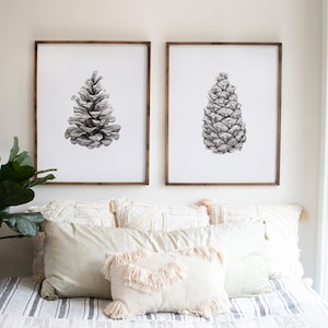 May include: Two black and white framed prints of pine cones on a white wall above a bed with a white and gray striped comforter and white pillows.