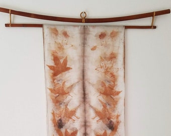 May include: A white silk scarf with a symmetrical design of brown leaves printed on it. The scarf is hanging from a wooden rod.