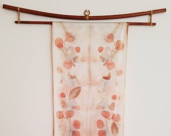 May include: A white silk scarf with a symmetrical design of pressed leaves and flowers in shades of brown and orange. The scarf is hanging from a wooden rod.