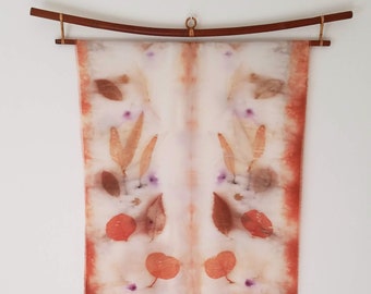 May include: A white silk scarf with a symmetrical design of orange and brown leaves and purple flowers. The scarf is hanging on a wooden rod.