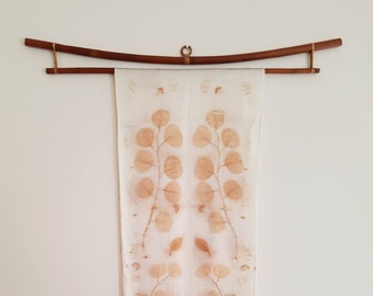 May include: A white silk scarf with a brown leaf pattern hanging on a wooden rod. The scarf has a symmetrical design with leaves on both sides.