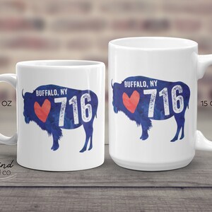 May include: Two white ceramic mugs with a blue and red design. The design features a silhouette of a buffalo with the words "BUFFALO, NY" and "716" on it. The smaller mug is 11 ounces and the larger mug is 15 ounces.