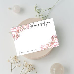 Op de afbeelding: Een witte kaart met de tekst "Memories of you" in sierlijke letters. De kaart is versierd met roze bloemaccenten in aquarel. De onderkant van de kaart heeft het woord "Love." gevolgd door een stippellijn voor een handtekening.
