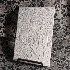 May include: A rectangular, white, textured art piece featuring a relief pattern of various leaves. The artwork is displayed on a dark metal stand, showcasing the intricate details of the leaf imprints.