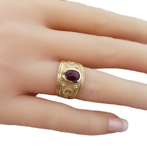 May include: A gold ring with an oval-shaped red gemstone set in the center. The band is decorated with a swirling pattern and small beads.