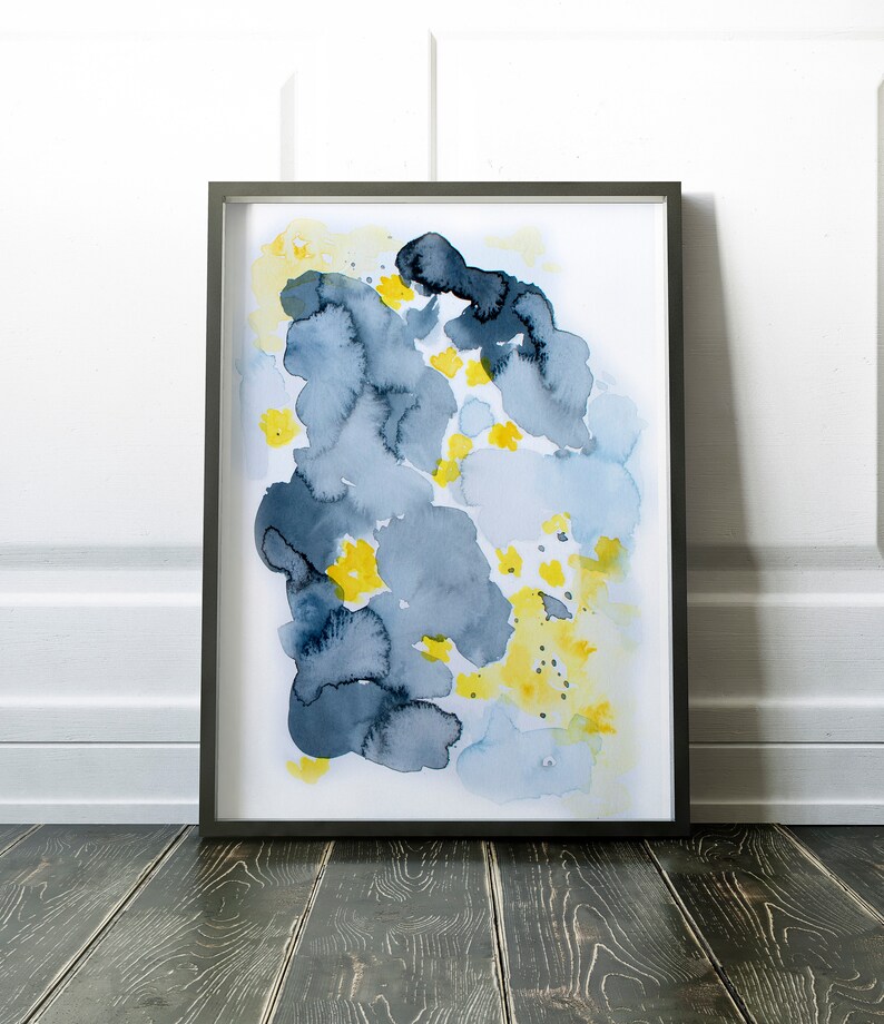 May include: Abstract watercolor art print in a black frame. The artwork features blue and gray cloud-like shapes with yellow floral accents on a white background. The framed art is displayed on a dark wooden floor.