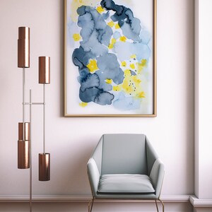 May include: An abstract watercolor painting in a gold frame, featuring blue, yellow, and dark blue shapes. A light blue armchair with a gold frame sits below the artwork. A modern floor lamp with copper shades is to the left.