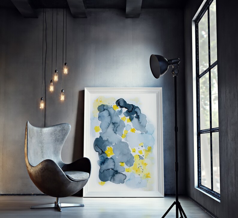 May include: An interior design scene featuring a gray leather egg chair, a framed abstract watercolor painting with blue and yellow hues, and a black floor lamp. Edison bulb pendant lights hang from the ceiling, and a large window is visible.