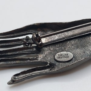 May include: A detailed, antique-style brooch in the shape of a hand, cast in a dark, metallic gray. The brooch features intricate detailing of the palm and fingers. The text "THELMA DEUTSCH" is visible on the back.
