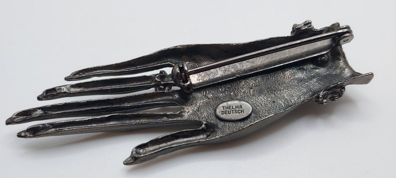 May include: A detailed, antique-style brooch in the shape of a hand, cast in a dark, metallic gray. The brooch features intricate detailing of the palm and fingers. The text "THELMA DEUTSCH" is visible on the back.