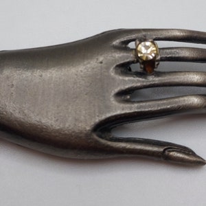 May include: A silver-toned hand-shaped brooch with two clear, faceted gemstones. One gemstone is a large, rectangular shape on the wrist, and the other is a smaller, round stone set in a ring on the middle finger. The brooch has a vintage aesthetic.