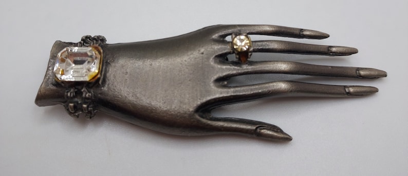 May include: A silver-toned hand-shaped brooch with two clear, faceted gemstones. One gemstone is a large, rectangular shape on the wrist, and the other is a smaller, round stone set in a ring on the middle finger. The brooch has a vintage aesthetic.