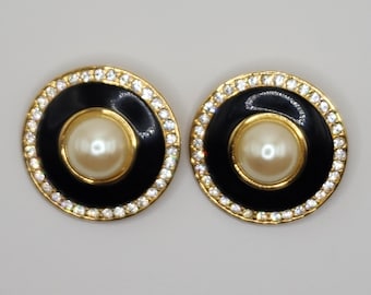 Könnte beinhalten: Ein Paar goldfarbene Ohrringe mit schwarzem Emaille und einem Perlmutt-Imitat in der Mitte. Die Ohrringe sind von klaren Strasssteinen umgeben.