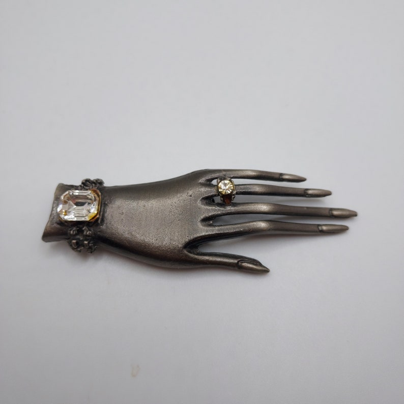 May include: A dark gray metal hand-shaped brooch with a ring on the middle finger and a bracelet on the wrist. The ring and bracelet feature clear, rectangular and round-cut gemstones.