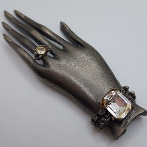 May include: A silver-toned hand-shaped brooch with two clear, sparkling gemstone accents. One gemstone is set in a ring on a finger, and the other is a large, rectangular stone on the wrist. The brooch has a vintage aesthetic.