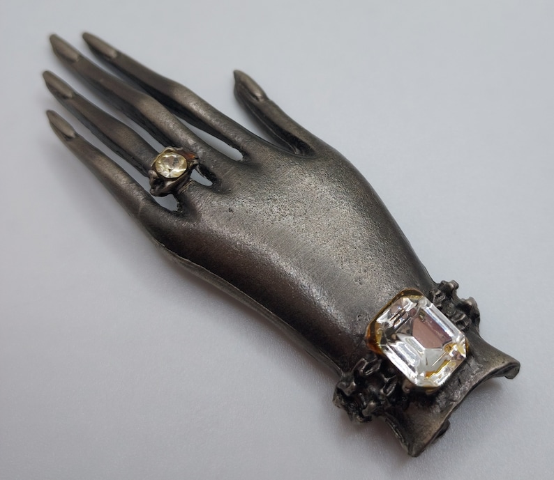 May include: A silver-toned hand-shaped brooch with two clear, sparkling gemstone accents. One gemstone is set in a ring on a finger, and the other is a large, rectangular stone on the wrist. The brooch has a vintage aesthetic.