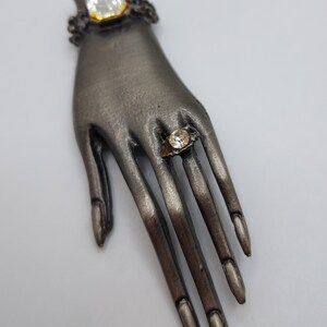 May include: A silver-toned hand-shaped brooch with two clear, square-cut gemstones. One gemstone is set at the wrist, and the other is on a finger, resembling a ring. The brooch has detailed finger and nail features.