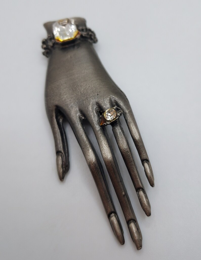 May include: A silver-toned hand-shaped brooch with two clear, square-cut gemstones. One gemstone is set at the wrist, and the other is on a finger, resembling a ring. The brooch has detailed finger and nail features.