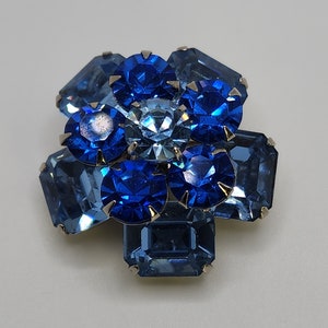 May include: A blue and light blue rhinestone brooch in the shape of a flower. The brooch is made of faceted rhinestones set in silver tone metal.