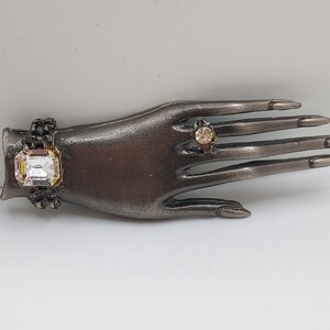 May include: A decorative hand-shaped jewelry holder in a dark metallic finish. It features a large, square-cut clear crystal bracelet and a small ring with a round, amber-colored stone. The fingers are long and slender.