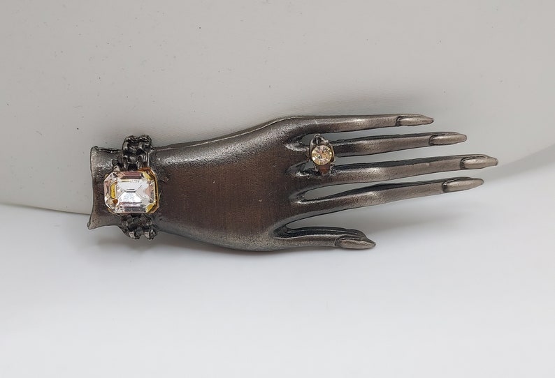 May include: A decorative hand-shaped jewelry holder in a dark metallic finish. It features a large, square-cut clear crystal bracelet and a small ring with a round, amber-colored stone. The fingers are long and slender.
