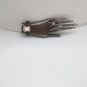 May include: A bronze-toned hand-shaped jewelry display featuring a bracelet with a large, clear, rectangular stone and a ring with a smaller, round stone. The hand has long, dark nails.