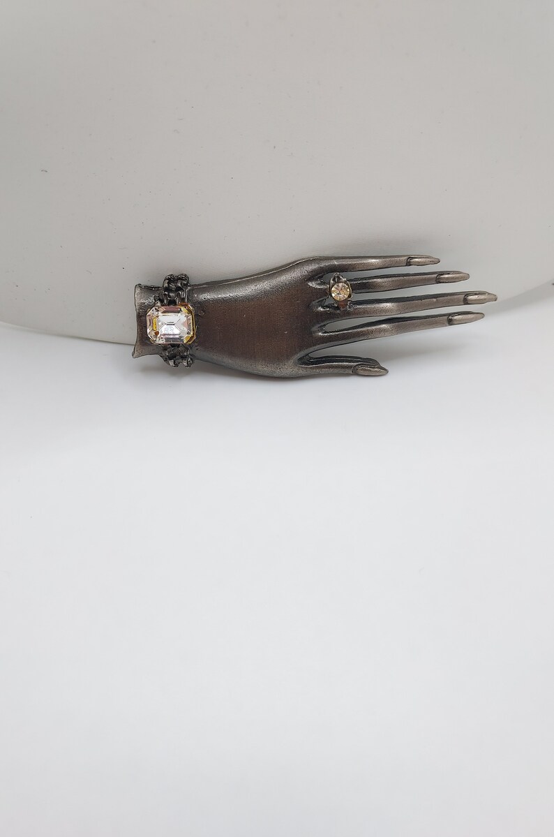 May include: A bronze-toned hand-shaped jewelry display featuring a bracelet with a large, clear, rectangular stone and a ring with a smaller, round stone. The hand has long, dark nails.