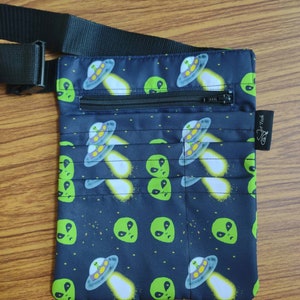 May include: A black fanny pack with a zipper closure and an adjustable strap. The fanny pack has a repeating pattern of green alien faces and flying saucers on a black background.