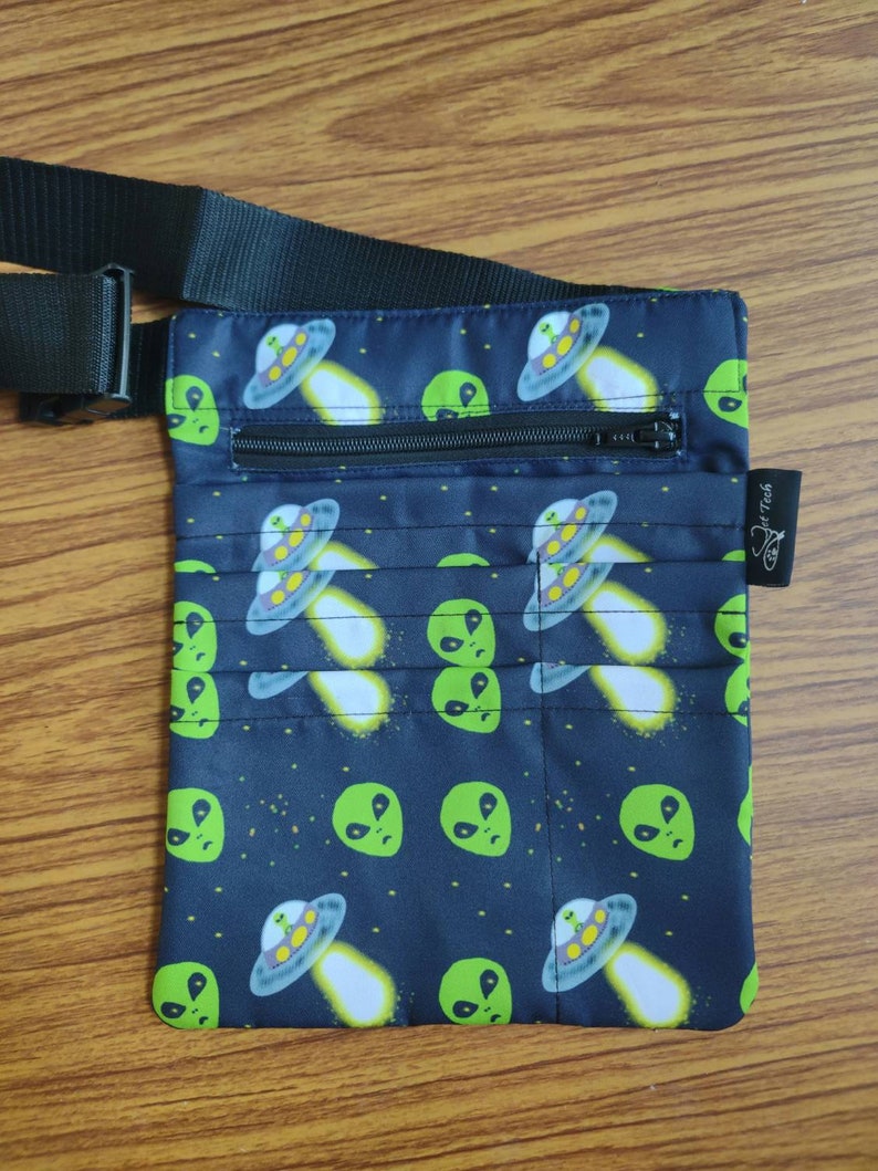 May include: A black fanny pack with a zipper closure and an adjustable strap. The fanny pack has a repeating pattern of green alien faces and flying saucers on a black background.