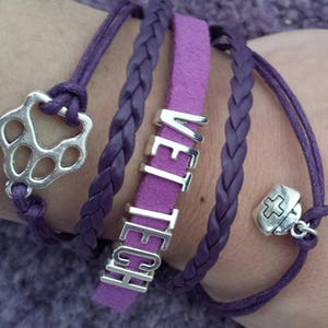 Op de afbeelding: Paarse gevlochten en su&egrave;de armbanden met zilveren bedels, waaronder een pootprint, een kruis en het woord "VET TECH".
