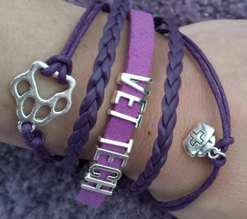 Op de afbeelding: Paarse gevlochten en su&egrave;de armbanden met zilveren bedels, waaronder een pootprint, een kruis en het woord "VET TECH".
