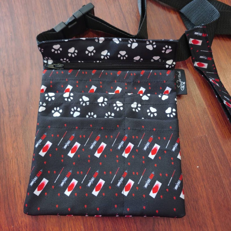 May include: Black fabric utility belt with multiple pockets and a zipper closure. The belt has a paw print pattern on the top flap and a red syringe pattern on the bottom. The belt has a black strap with a buckle.