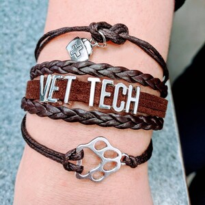 Op de afbeelding: Een bruin leren armband met de woorden "VET TECH" erop. De armband heeft een zilveren pootprintbedel en een zilveren bedel met een kruis.
