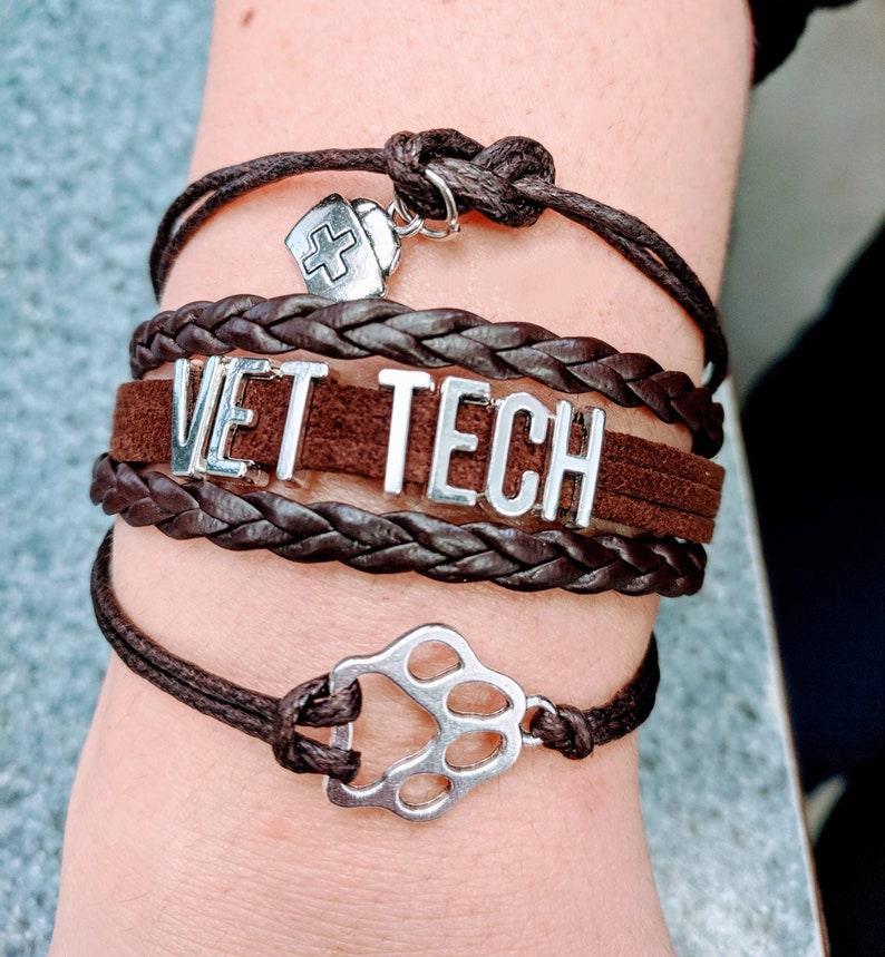 Op de afbeelding: Een bruin leren armband met de woorden "VET TECH" erop. De armband heeft een zilveren pootprintbedel en een zilveren bedel met een kruis.