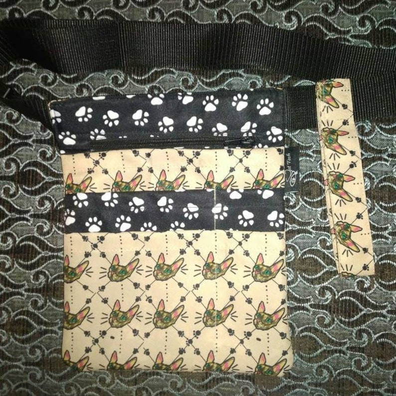 May include: A black and tan fabric bag with a zippered pocket and a paw print design. The bag has a black adjustable strap and a matching fabric pouch with a cat face design. The bag is labeled "Cat Tech".