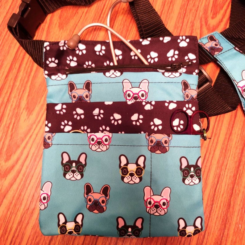 May include: A blue and black fabric utility bag with a zipper closure and multiple pockets. The bag features a repeating pattern of French Bulldogs wearing glasses. The bag has a black strap and a silver stethoscope.