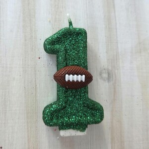 May include: A number one shaped birthday candle covered in green glitter. The candle features a brown textured American football with white laces attached to the center of the number. A small white wick extends from the top of the green glittered candle.