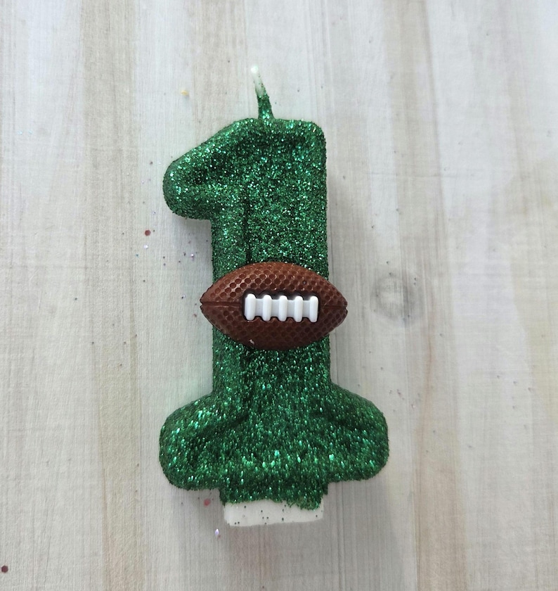 May include: A number one shaped birthday candle covered in green glitter. The candle features a brown textured American football with white laces attached to the center of the number. A small white wick extends from the top of the green glittered candle.