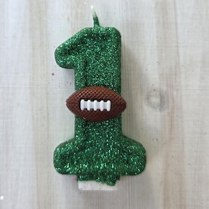 May include: A number one shaped birthday candle covered in green glitter. A small brown football with white laces is attached to the center of the candle. The candle features a single white wick at the top and a flat base for standing upright.